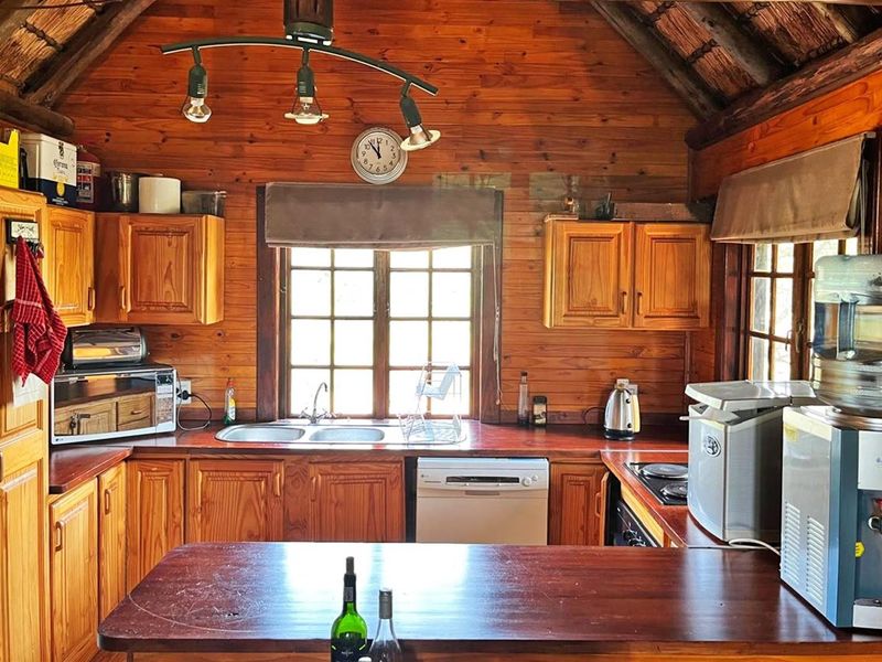 A rustic kitchen featuring timber wall cladding, wooden cabinetry, a double bowl sink, and integrated appliances under a traditional thatch roof.