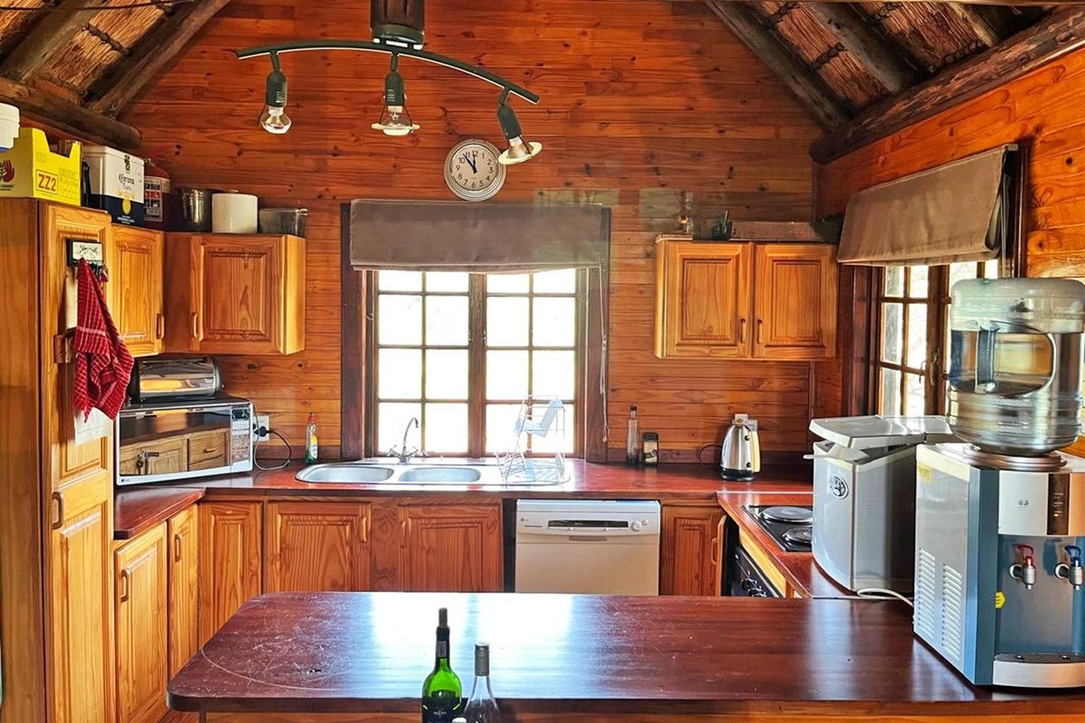 A rustic kitchen featuring timber wall cladding, wooden cabinetry, a double bowl sink, and integrated appliances under a traditional thatch roof.