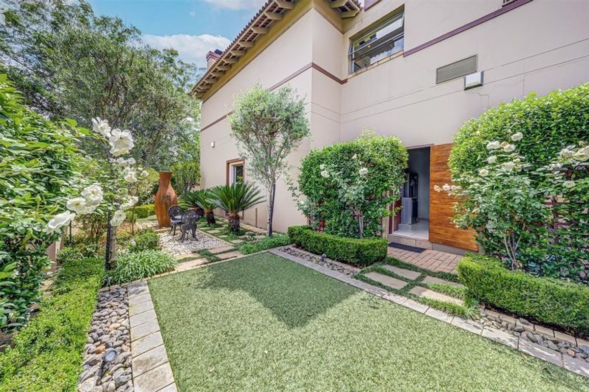 A well-maintained rear garden featuring artificial lawn, stone paving, mature landscaping, and direct access to the home via a timber door.