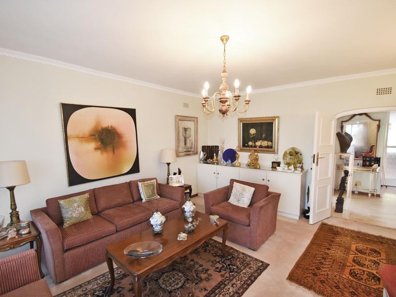 A well-maintained living room featuring classic ceiling cornicing, a central chandelier, and built-in cabinetry, leading through an archway to an adjacent space.