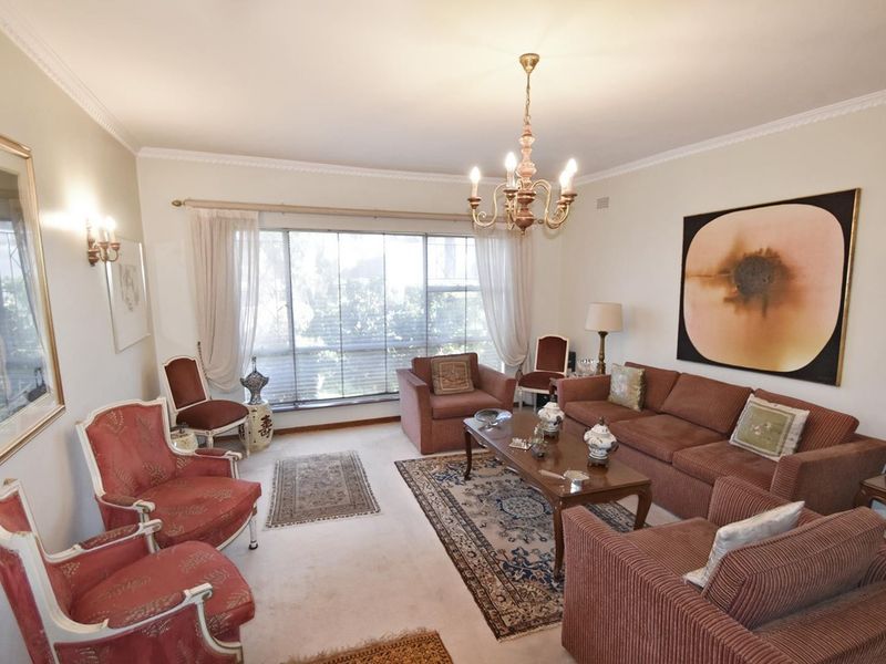 A spacious living room featuring classic ceiling cornicing, a large window allowing natural light, and a central chandelier.