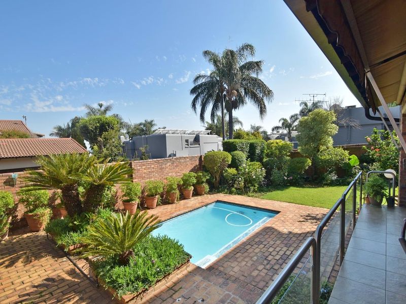 A private backyard featuring a swimming pool, brick-paved patio area, and a covered veranda with a glass balustrade overlooking the garden.