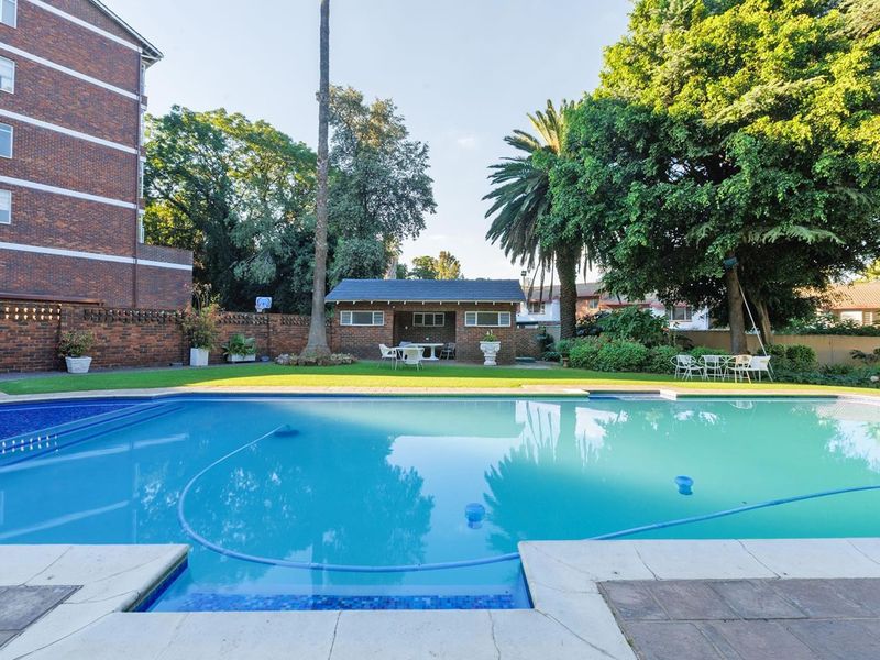A well-maintained outdoor pool area featuring a clear swimming pool, paved surround, and a brick outbuilding set within a landscaped garden.