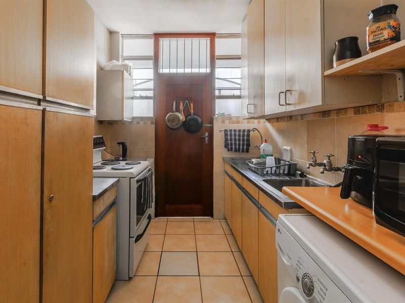 A compact, dated kitchen featuring original cabinetry, tiled flooring, a stainless steel sink, and a freestanding stove with direct access to an exterior door.