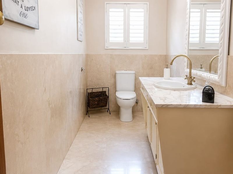A well-maintained bathroom featuring a marble-topped vanity with brass fixtures, tiled walls, and plantation shutters for natural light.