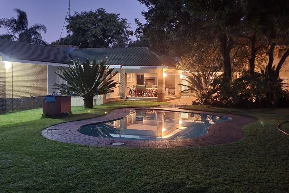 Night view of the rear exterior featuring a swimming pool with brick coping, a covered patio area, and a well-maintained garden.