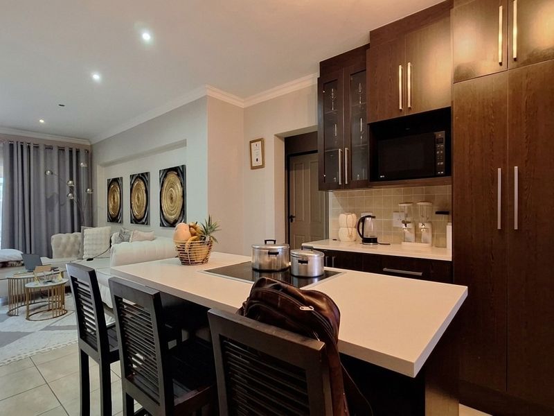 Modern open-plan kitchen and living area featuring dark wood cabinetry, stone countertops, integrated appliances, and recessed lighting, flowing into a bright lounge space.