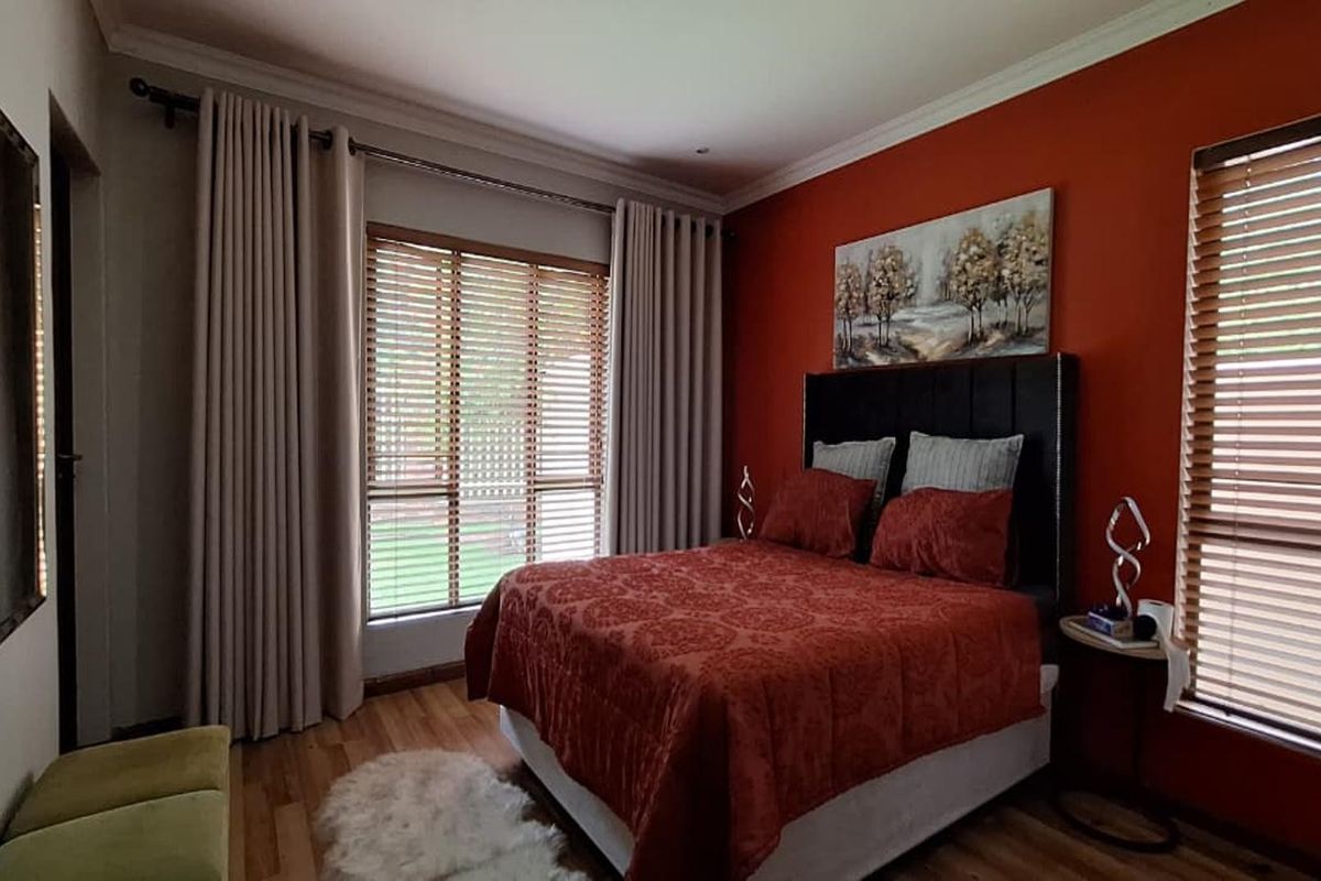 A well-maintained bedroom featuring warm-toned walls, wooden flooring, and large windows with fitted venetian blinds and curtain tracks.