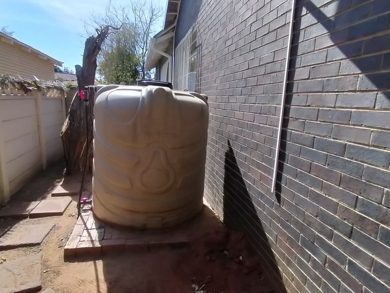 Side exterior area featuring a large plastic water tank positioned against a brick wall with paved ground and boundary fencing.