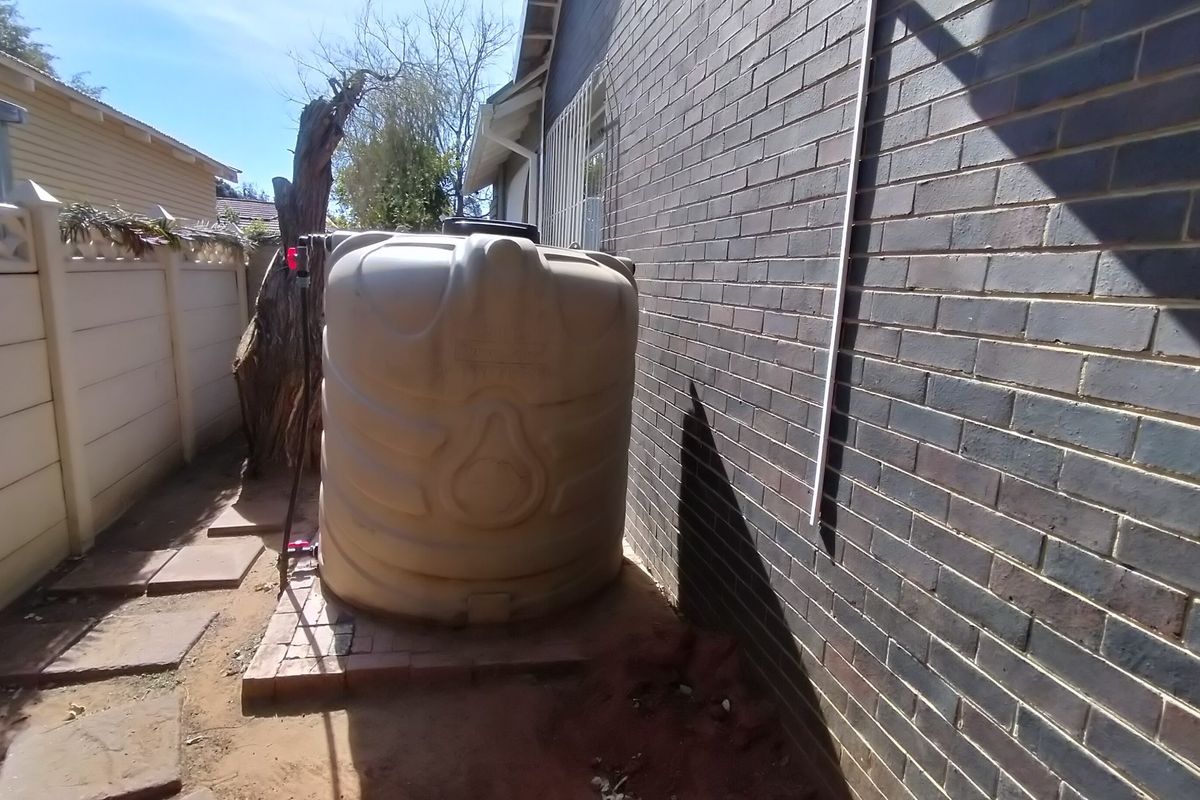 Side exterior area featuring a large plastic water tank positioned against a brick wall with paved ground and boundary fencing.
