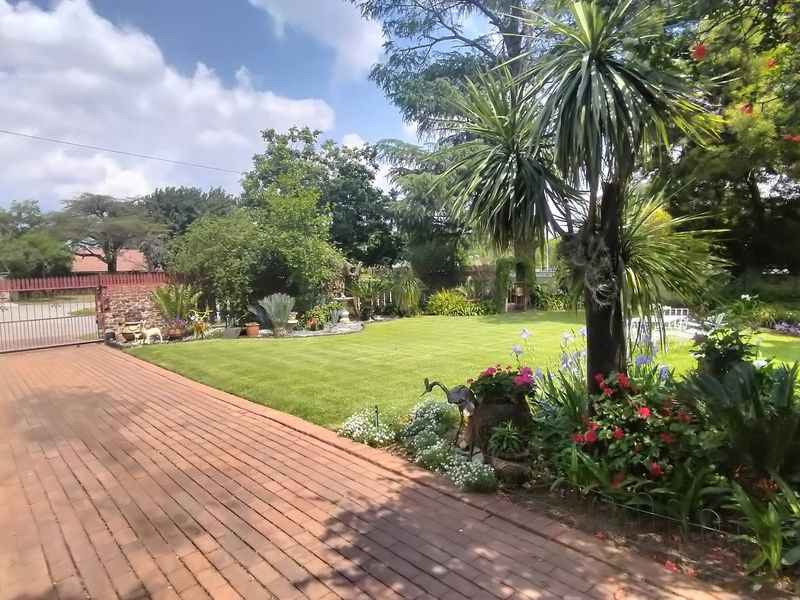 A well-maintained garden featuring a brick-paved driveway, a metal entrance gate, and established landscaping with a lush green lawn.
