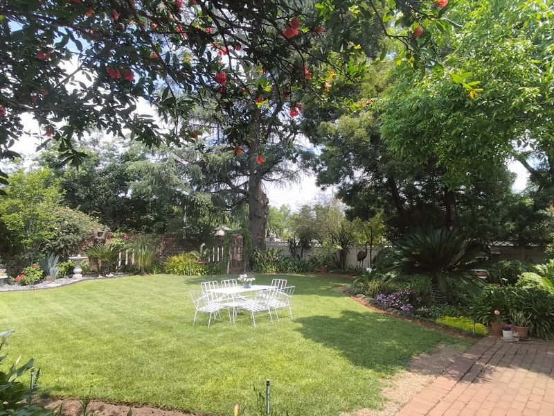 A well-maintained, lush green garden featuring mature trees, established landscaping, and a brick-paved patio area.