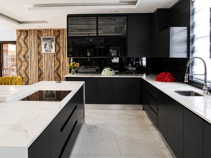 Modern kitchen featuring sleek black cabinetry, white stone countertops, a large central island with an integrated hob, and stylish decorative wall paneling.