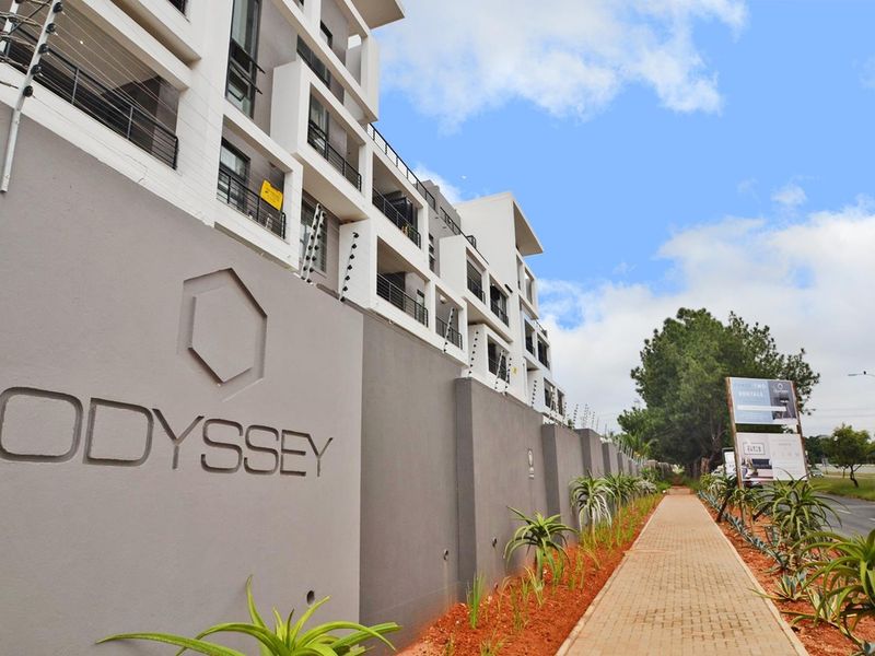 Modern apartment complex exterior featuring a paved walkway, landscaped garden beds, and secure perimeter wall with electric fencing.