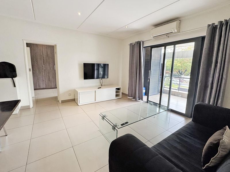 A bright living room featuring neutral tiled flooring, recessed lighting, a wall-mounted air conditioning unit, and sliding glass doors leading to a private balcony.