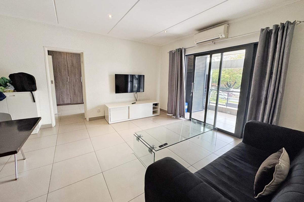 A bright living room featuring neutral tiled flooring, recessed lighting, a wall-mounted air conditioning unit, and sliding glass doors leading to a private balcony.