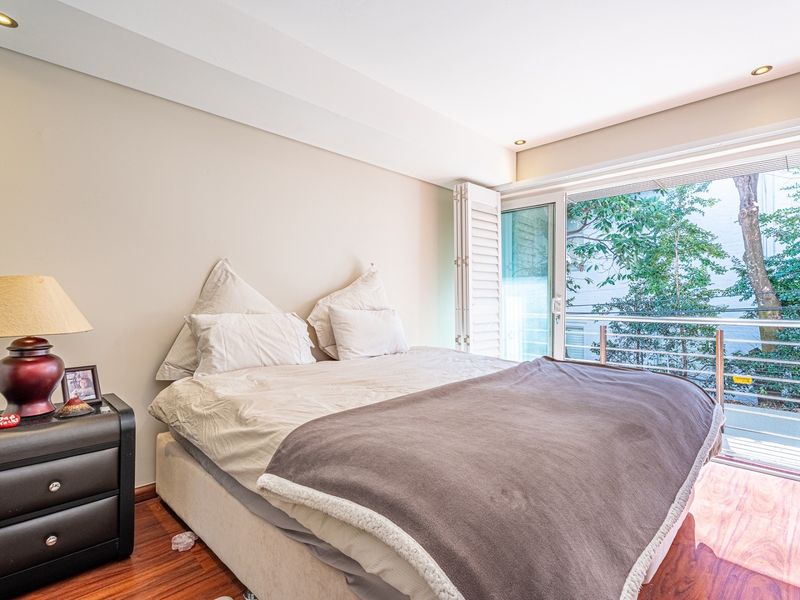 A bright bedroom featuring hardwood flooring, recessed lighting, and a sliding glass door with plantation shutters opening onto a balcony.