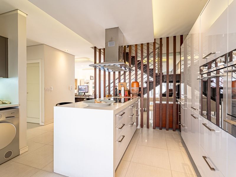 Modern kitchen featuring a central island with integrated hob, high-gloss cabinetry, built-in appliances, and a stylish timber slat divider adjacent to the staircase.