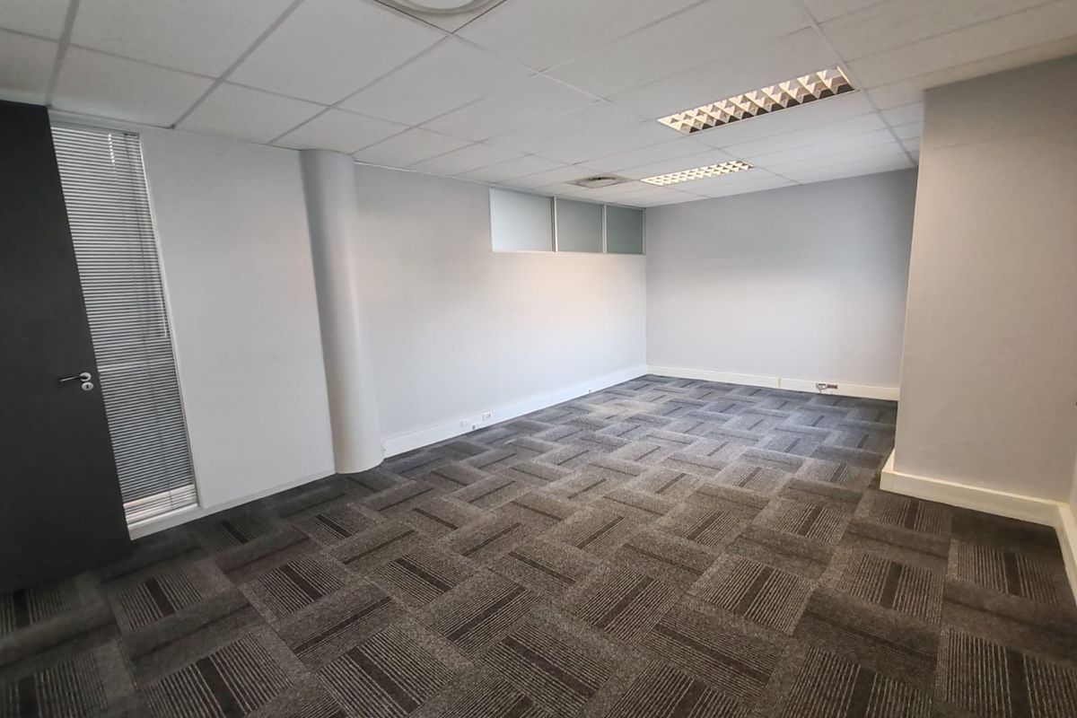 A clean, vacant office space featuring neutral grey walls, carpet tile flooring, and a suspended ceiling with integrated lighting.