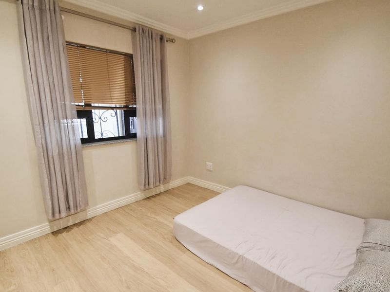 A bright and neutral bedroom featuring light laminate flooring, recessed downlighting, and a window fitted with security bars.