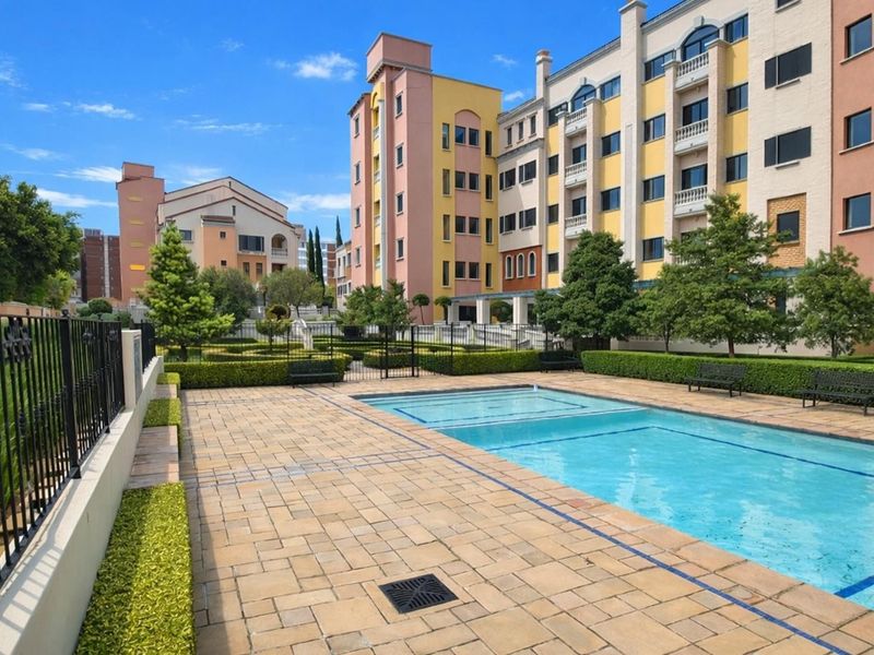 A well-maintained communal swimming pool area featuring paved decking, manicured hedges, and wrought iron fencing, set against a multi-story apartment complex.