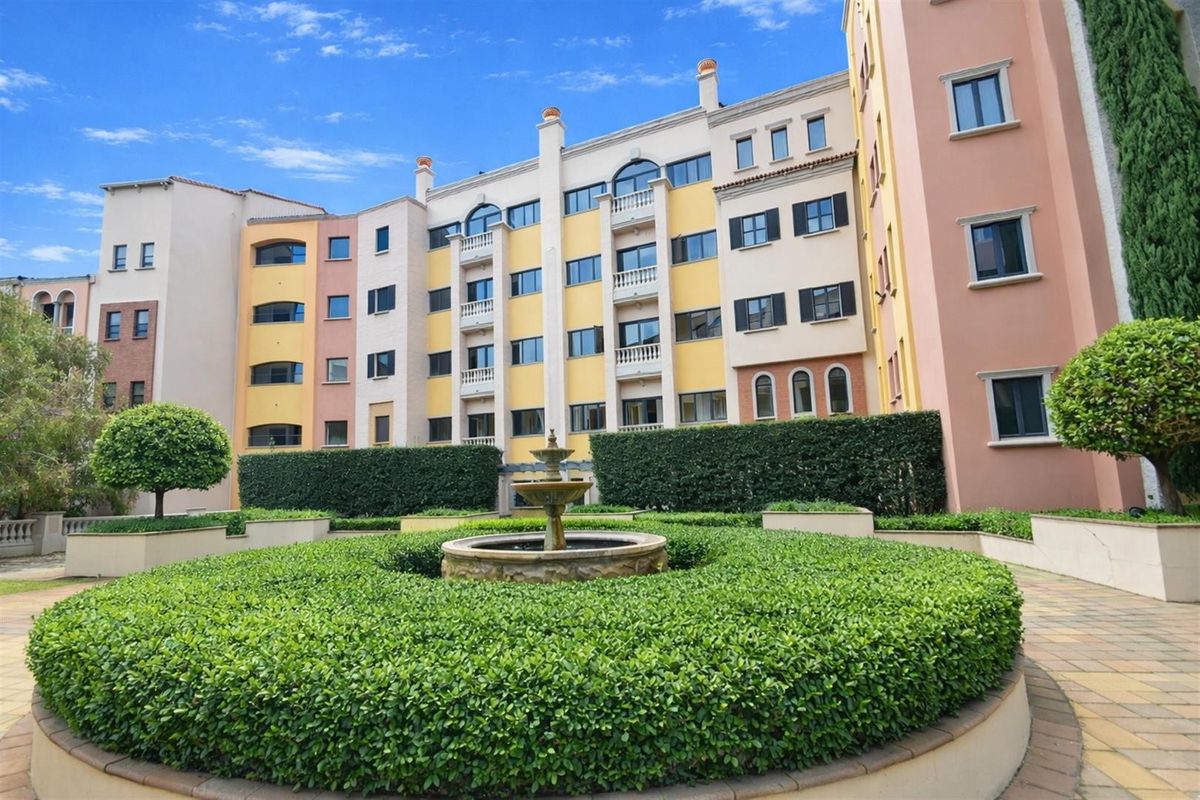 A well-maintained apartment complex courtyard featuring a central stone fountain, manicured hedges, and paved walkways surrounded by multi-story residential building facades.