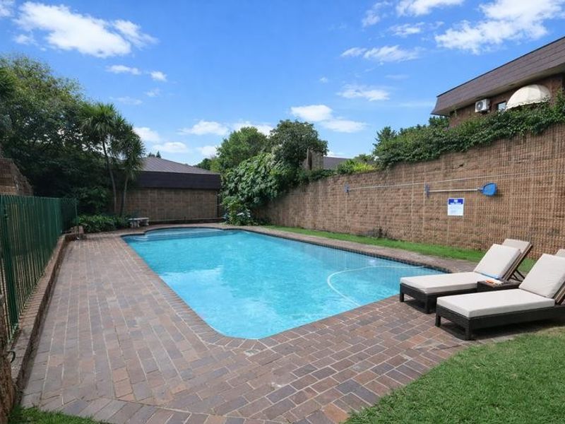 A well-maintained outdoor swimming pool area featuring brick paving, secure metal fencing, and high boundary walls for privacy.