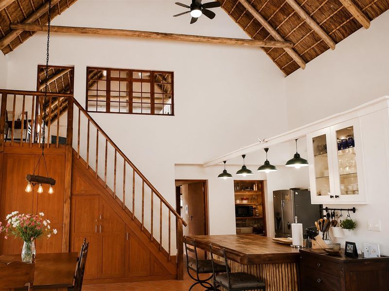 Spacious open-plan living area featuring a high vaulted thatch ceiling, exposed timber beams, a wooden staircase, and a well-appointed kitchen with a central island.