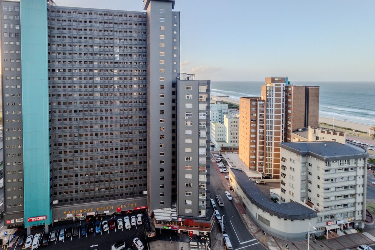 Aerial view of a high-rise apartment complex featuring ground-level commercial space and parking, situated near the coastline.