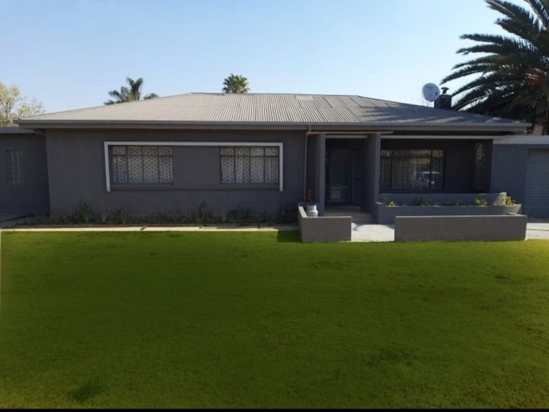 Front elevation of a single-story house featuring a grey painted facade, corrugated metal roof, and a well-maintained front lawn.