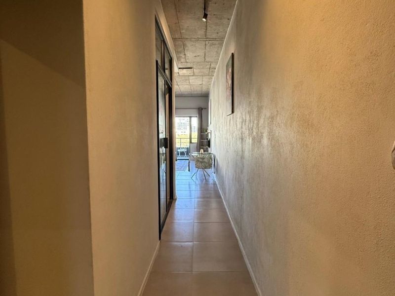 A modern hallway featuring an exposed concrete ceiling with track lighting and neutral tiled flooring, leading towards an open living area.