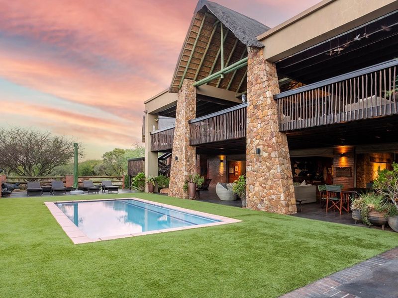 Rear view of a luxury residence featuring a swimming pool, artificial lawn, covered patio, and a distinctive thatched roof with stone pillar accents.