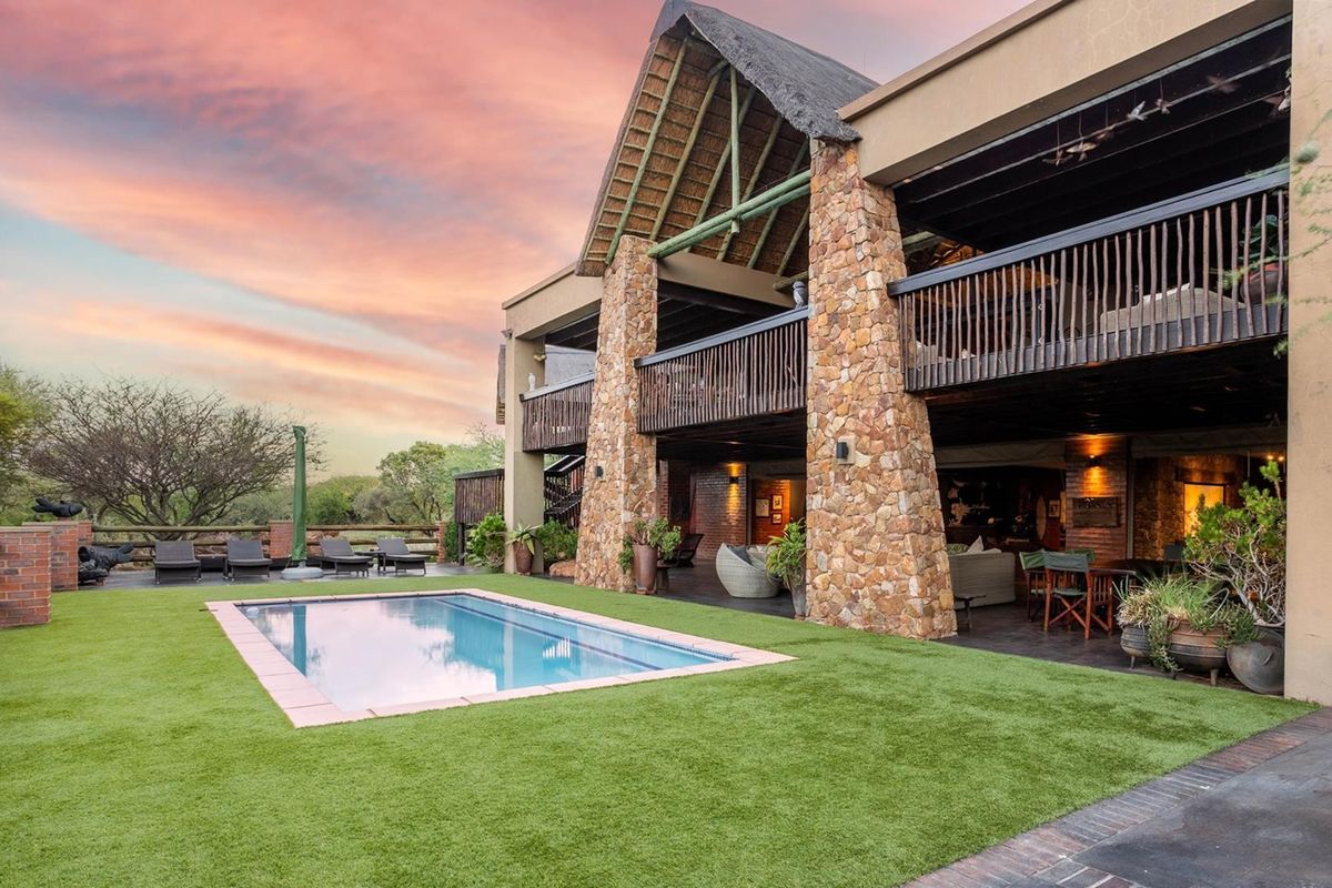 Rear view of a luxury residence featuring a swimming pool, artificial lawn, covered patio, and a distinctive thatched roof with stone pillar accents.