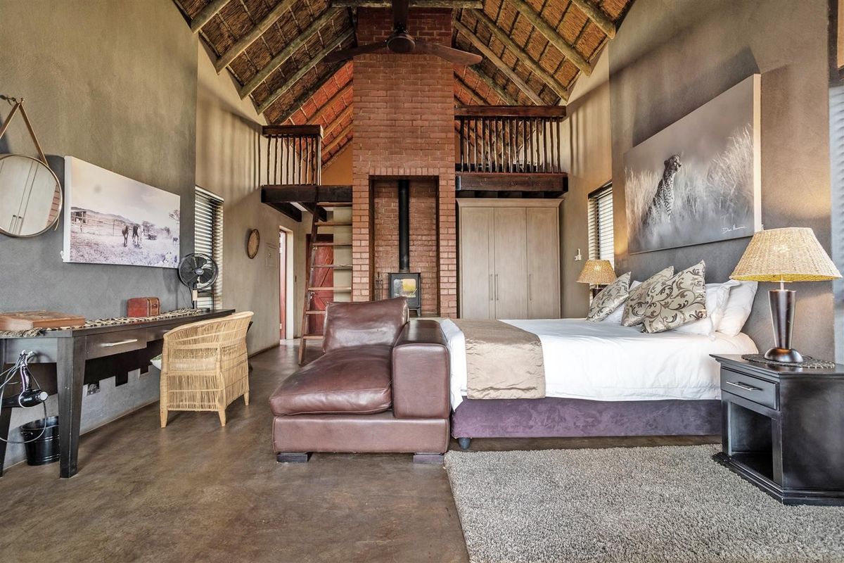 Spacious bedroom featuring a high exposed thatch ceiling, central brick fireplace with wood-burning stove, mezzanine loft area, and polished concrete flooring.