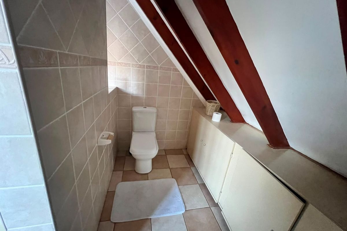 A compact bathroom featuring tiled finishes, a toilet, and built-in storage under the sloping roof with exposed timber rafters.