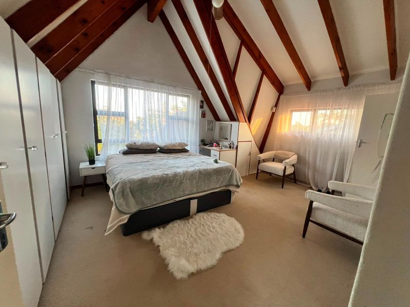 Spacious bedroom featuring exposed timber rafters, a sloped ceiling, built-in cupboards, and carpeted flooring with natural light from large windows.
