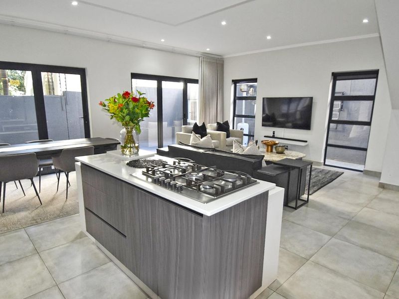 Modern open-plan living area featuring a central kitchen island with gas cooktop, tiled flooring, and large glass doors leading to the exterior.
