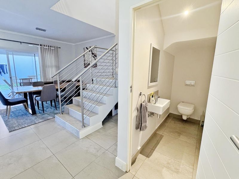 A bright hallway featuring a modern staircase with stainless steel railings, leading to an adjacent guest bathroom with wall-mounted fixtures and tiled flooring.