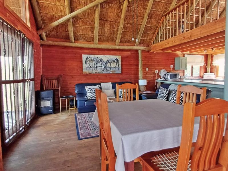 Open-plan living and dining area featuring rustic timber walls, exposed thatched roofing, and a mezzanine level, creating a warm, cabin-style atmosphere.