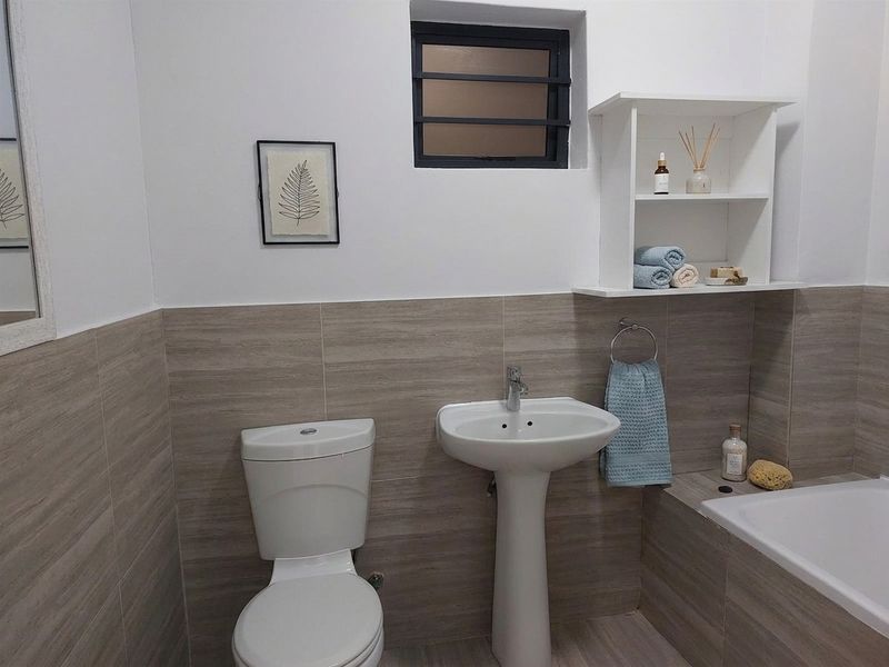 A functional bathroom featuring a pedestal basin, toilet, bathtub, and neutral wall tiling with built-in shelving.