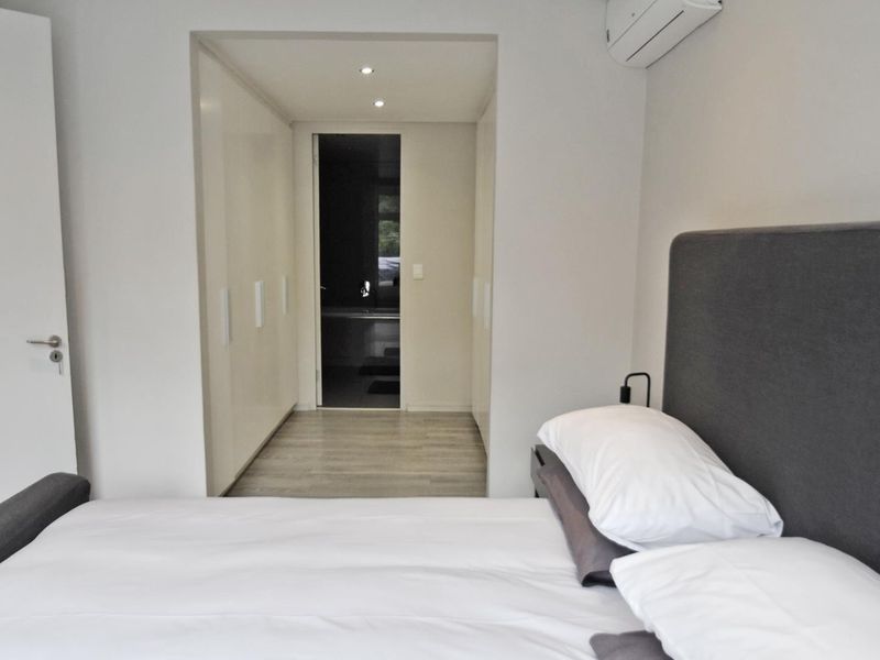 Modern bedroom featuring built-in cupboards, recessed lighting, and a wall-mounted air conditioning unit, leading into an adjoining bathroom area.