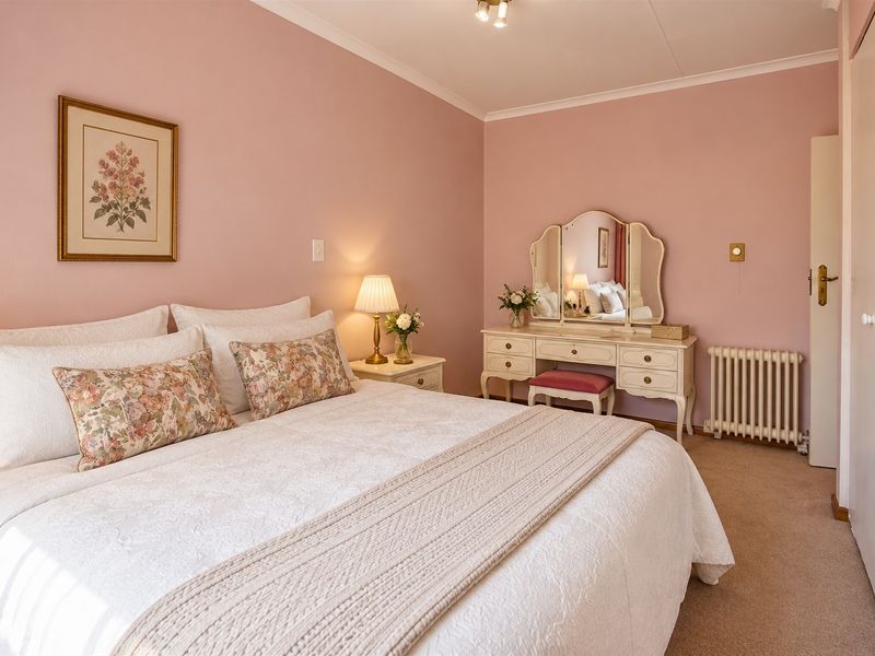 A well-maintained bedroom featuring soft pink walls, carpeted flooring, a built-in wardrobe, and a traditional radiator heating unit.
