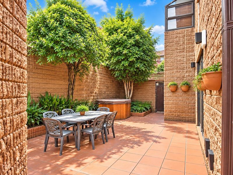 A private, paved outdoor patio area featuring brick boundary walls, established trees, and built-in garden beds, ideal for outdoor dining and relaxation.