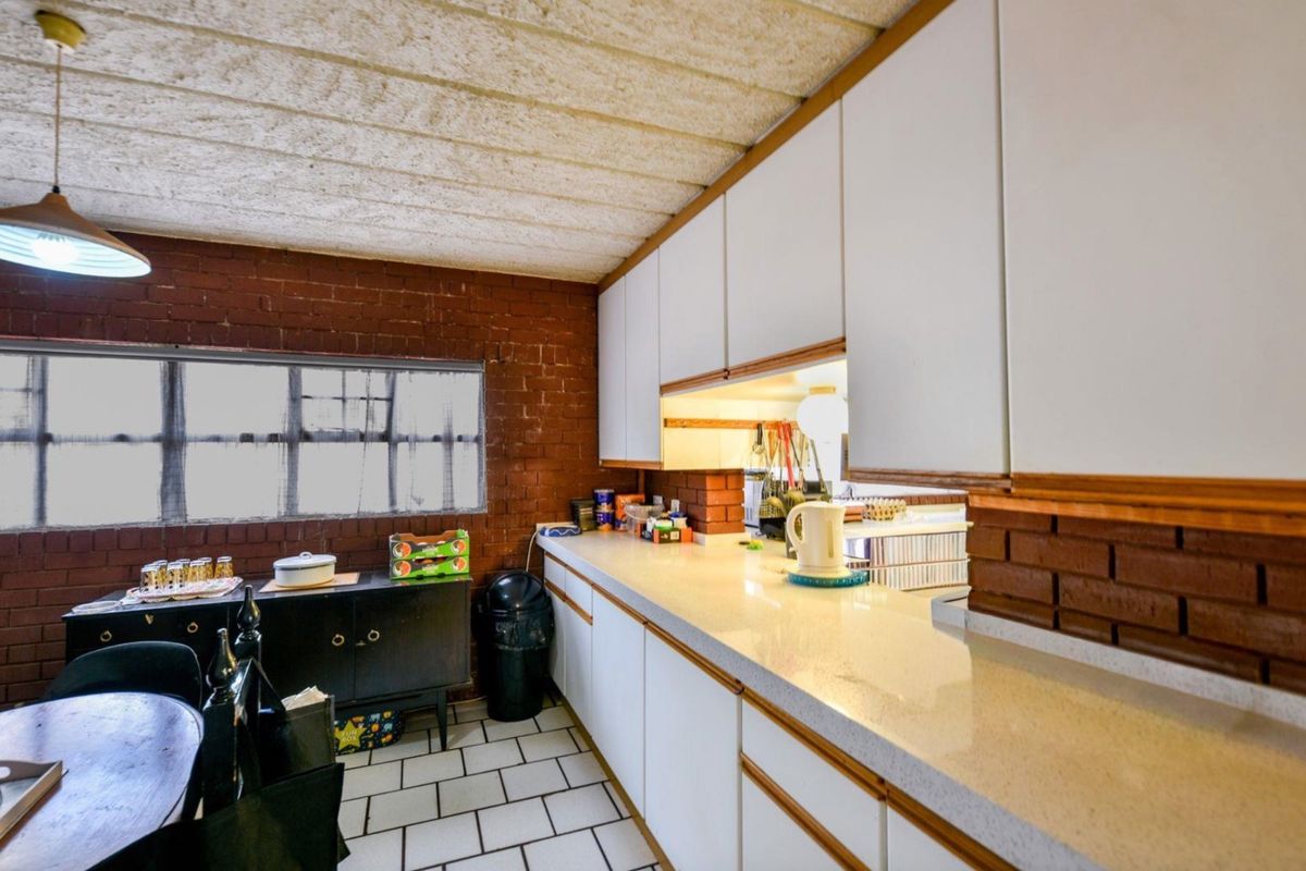 A dated kitchen featuring exposed brick walls, white cabinetry, stone-look countertops, and tiled flooring, offering significant potential for a modern renovation.