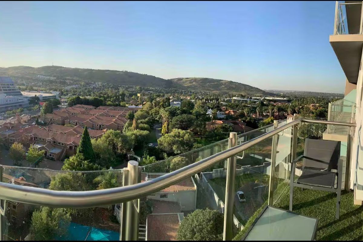 A modern balcony featuring a glass balustrade with a stainless steel handrail, offering expansive elevated views of the surrounding neighbourhood and hills.