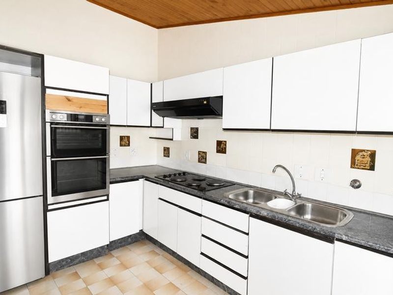 A functional kitchen featuring white cabinetry, built-in double oven, electric hob, and double sink with a tiled backsplash and timber ceiling.