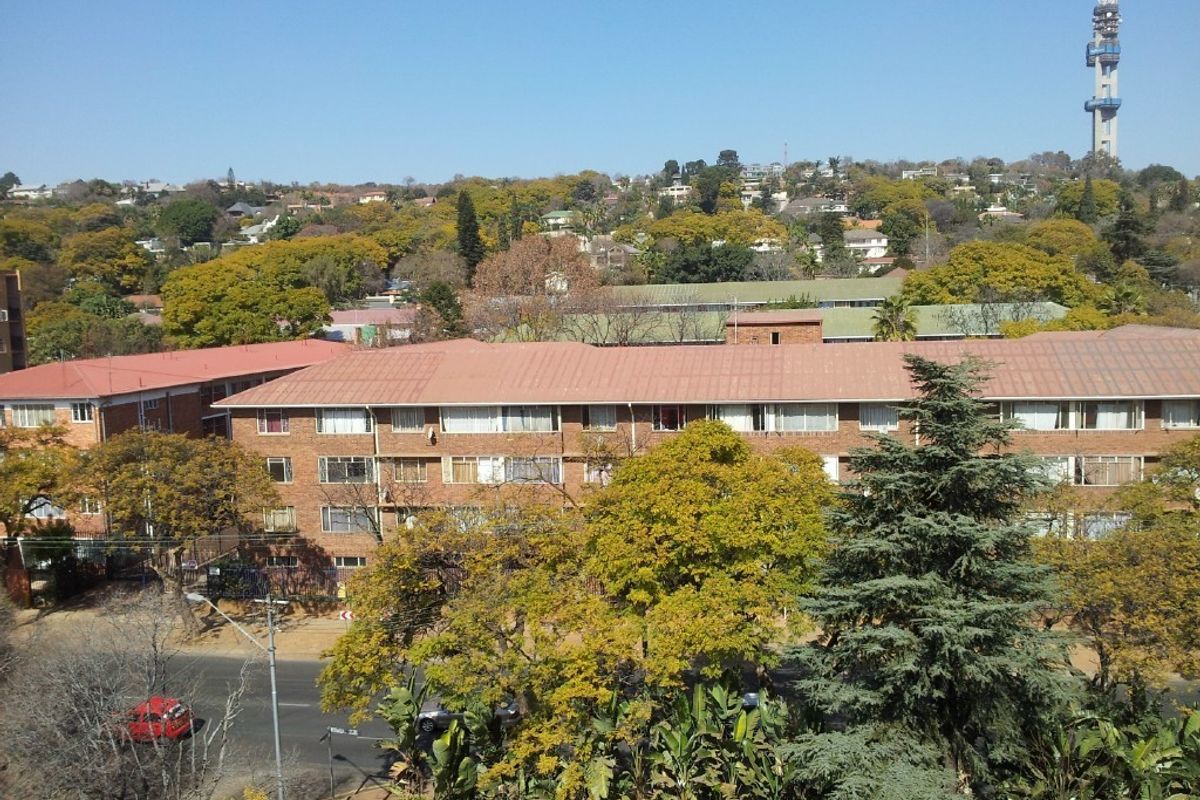 An elevated exterior view of a multi-story brick apartment complex surrounded by mature trees and urban landscape.