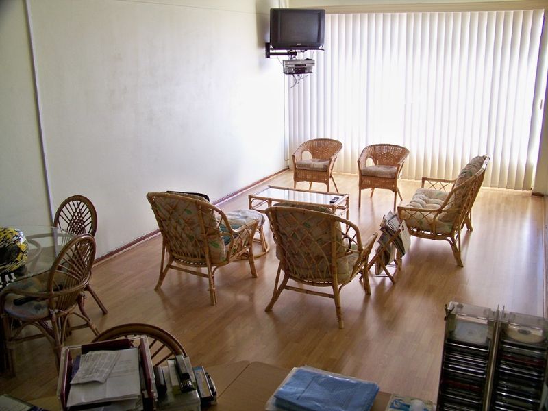 A living room area featuring laminate flooring and vertical blinds, currently furnished with casual seating and a wall-mounted television mount.