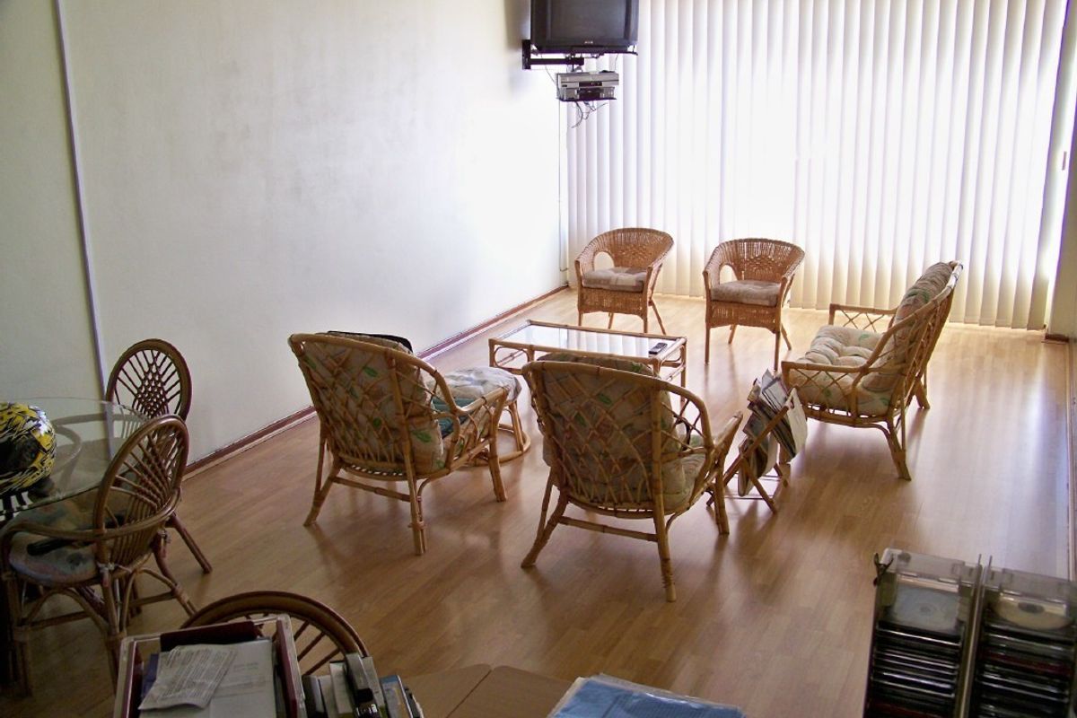 A living room area featuring laminate flooring and vertical blinds, currently furnished with casual seating and a wall-mounted television mount.