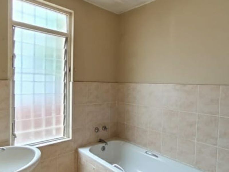 A functional bathroom featuring a bathtub, pedestal basin, and louvered window with neutral wall and tile finishes.